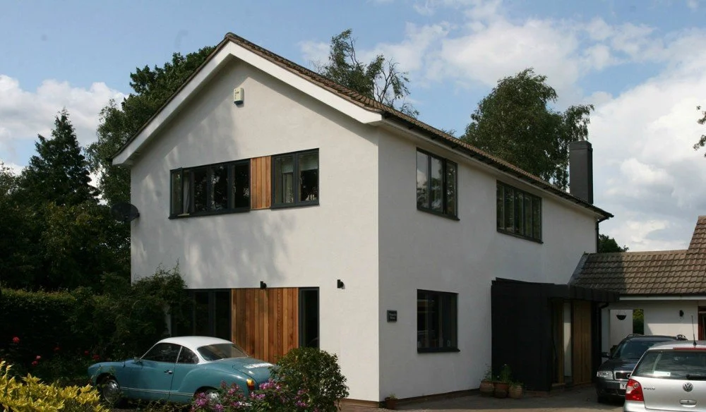 A modern, two-story house with white exterior walls, large windows, and a pitched roof with brown shingles. There is a vintage blue car parked on the driveway and several other modern cars nearby. The yard is landscaped with plants and bushes.