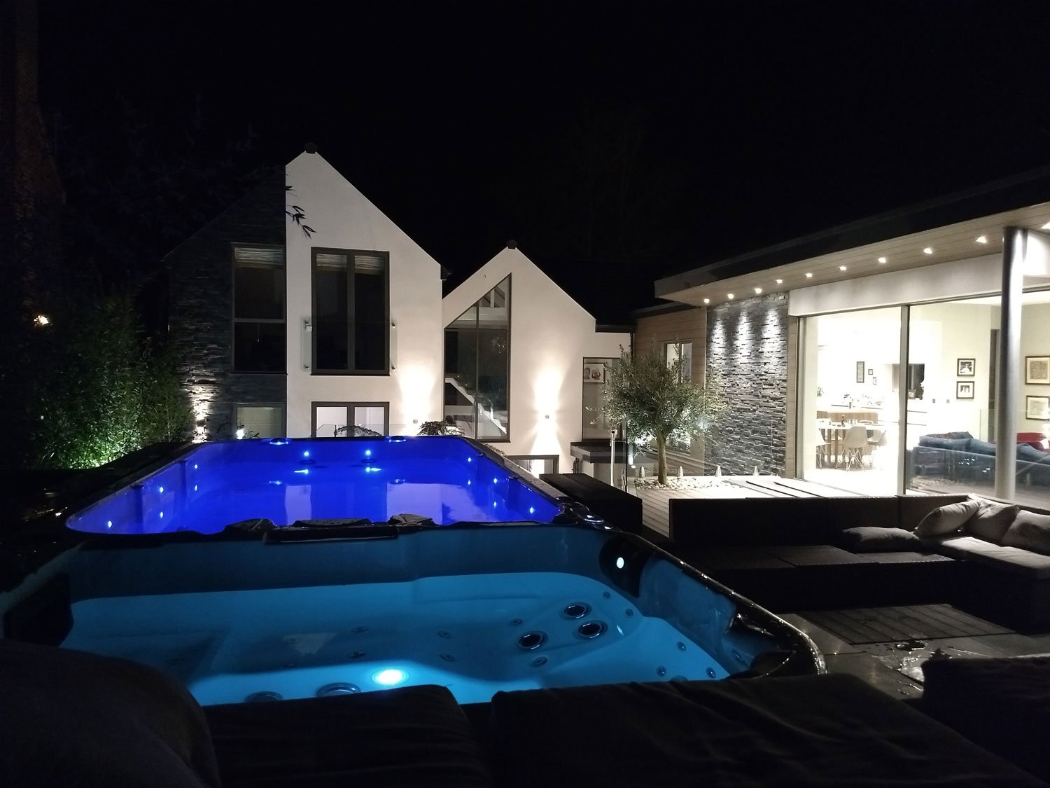 Night view of a modern house with large glass windows, outdoor seating area, illuminated pool with blue lighting, and Jacuzzi with blue underwater lights.