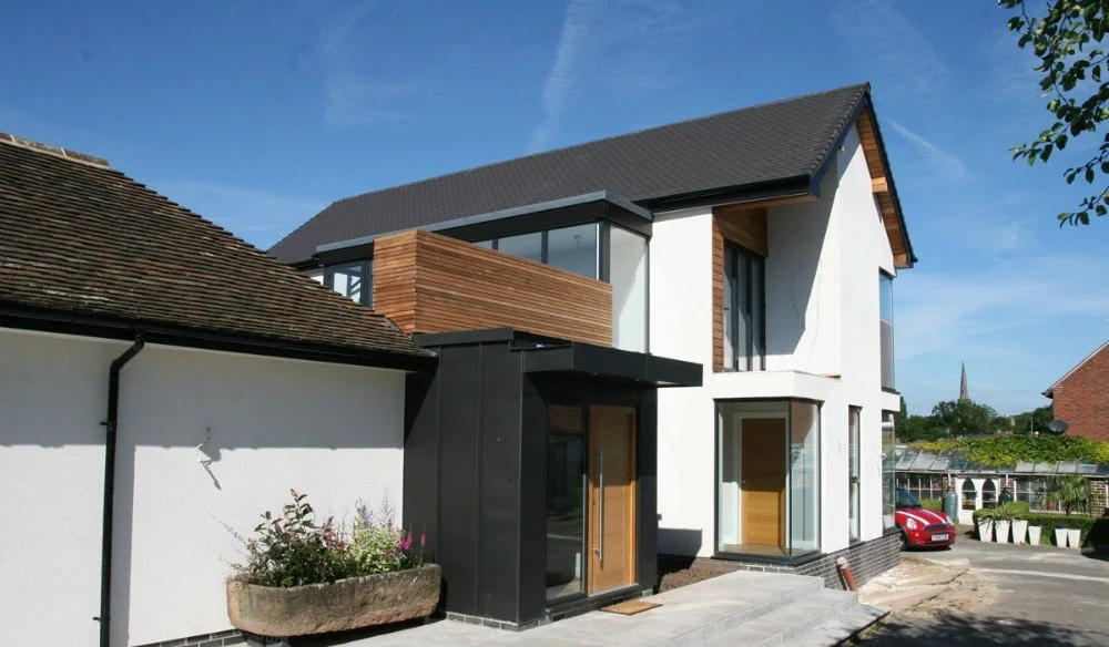 Modern two-story house with white walls, black accents, and wooden trim, with a car parked nearby and neighboring houses in the background.