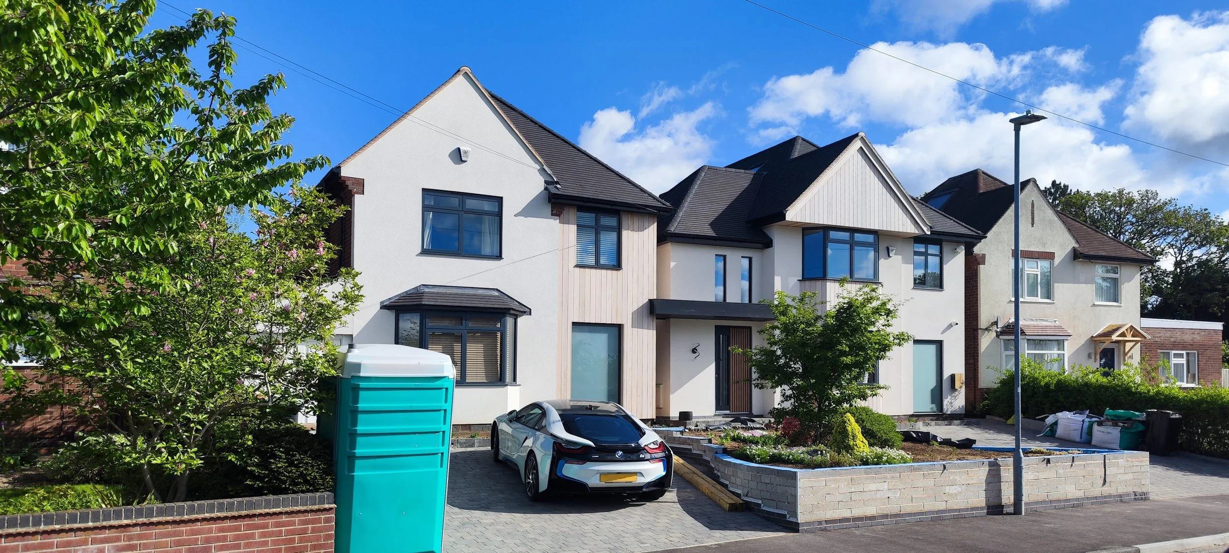 A modern white multi-story house with black window frames, surrounded by green trees and plants, under a partly cloudy blue sky. There is a black and white car parked in the driveway, and a green portable toilet is visible in the front yard, indicati