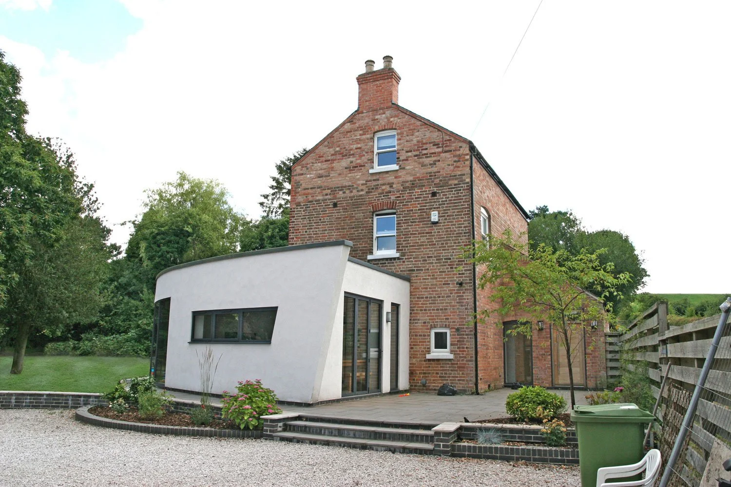 A multi-story brick house with a modern white extension, surrounded by a garden with trees, plants, and a gravel yard, with a wooden fence on the right side.