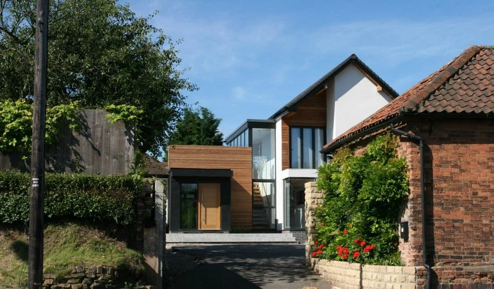 Modern two-story house with a mix of wood, white, and glass exterior, situated between older brick buildings, surrounded by greenery and trees.