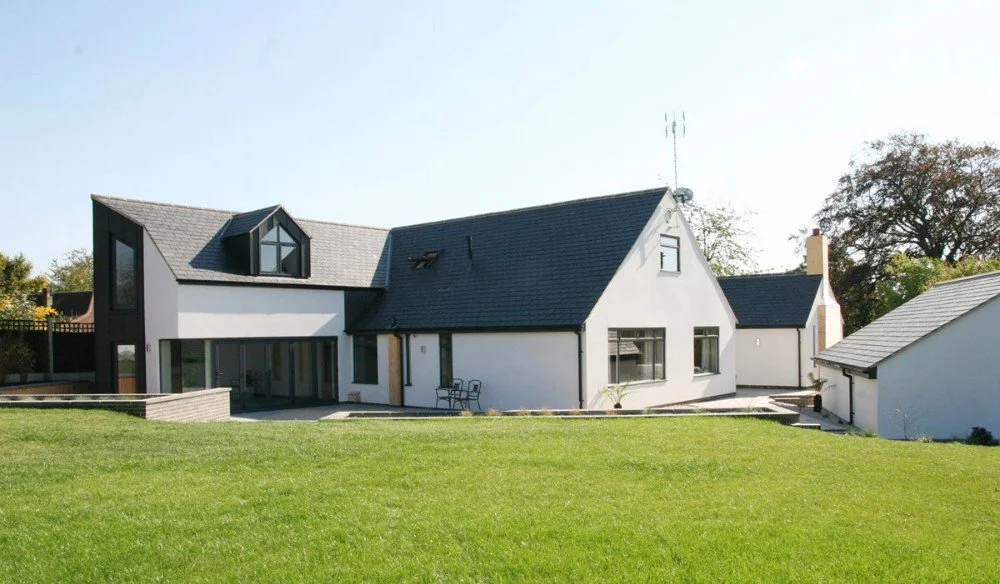 Modern house with a white exterior, dark gray roof, and large windows, set in a well-maintained lawn with trees in the background.