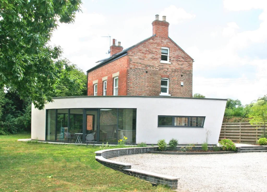 A modern white extension with large glass doors and a long horizontal window attached to a traditional red brick house, with a gravel driveway and small green plants in front, surrounded by trees.