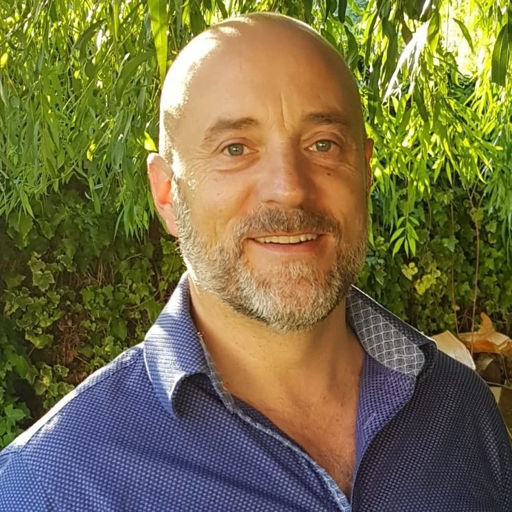 A smiling man with a beard and bald head outdoors, with green foliage in the background.