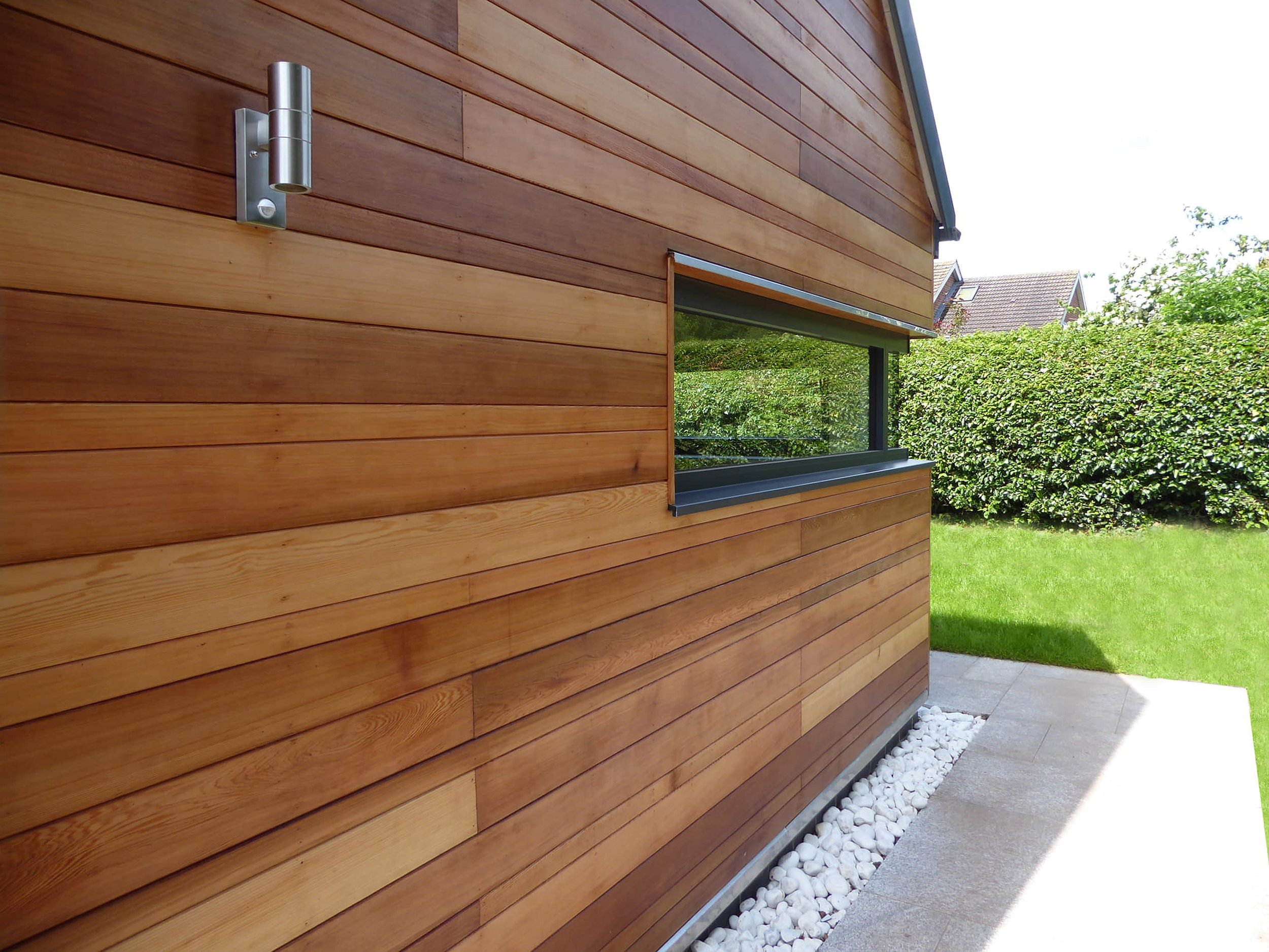 Exterior view of a modern house with horizontal wooden siding, a window with a gray frame, and a metal wall-mounted light fixture. There are bushes and a lawn in the background.