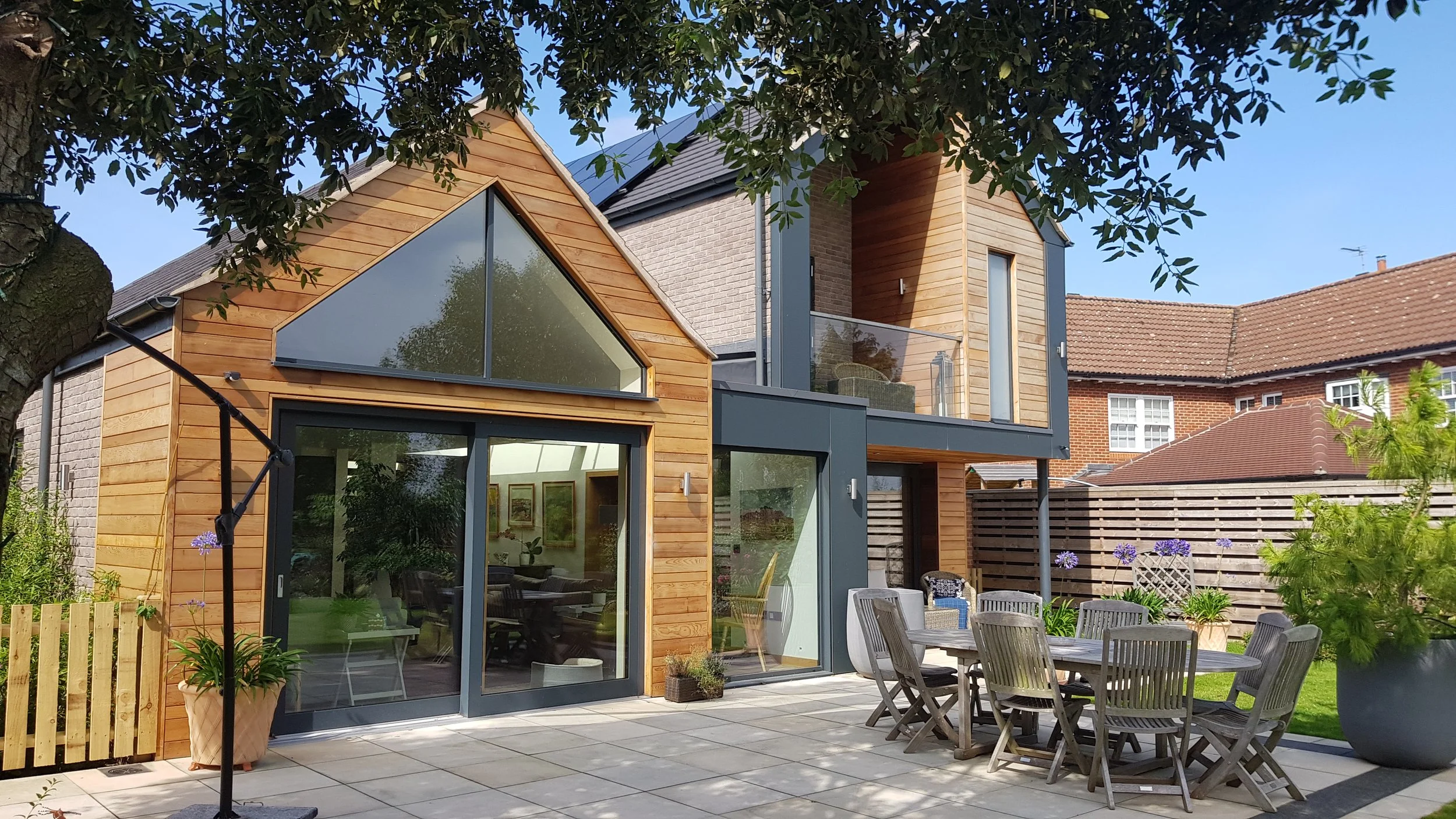 Modern two-story house with large glass windows and wooden siding, surrounded by a patio with outdoor furniture and potted plants, with neighboring houses visible in the background.