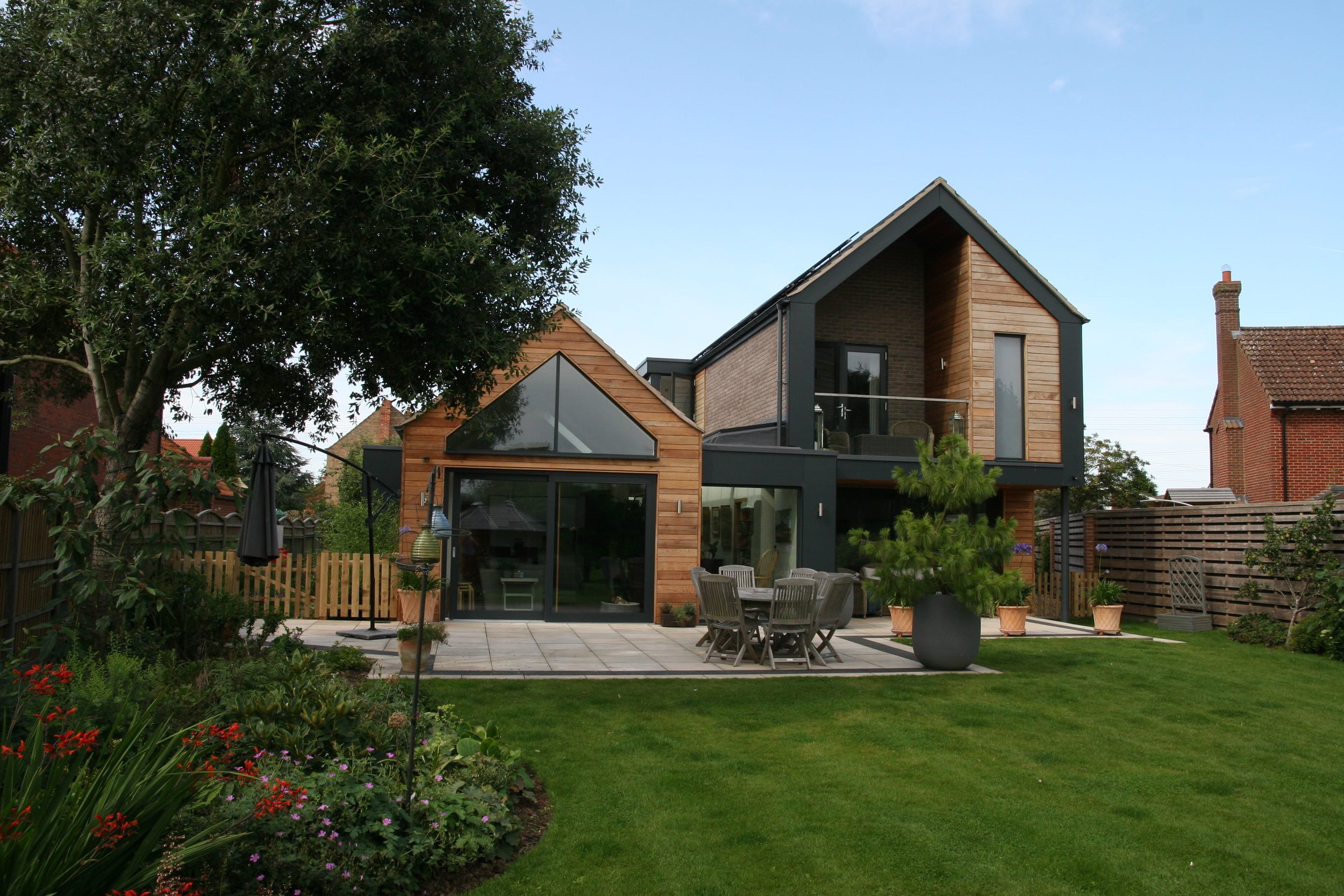 Modern house with a outdoor patio, surrounded by a garden and plants, with a large tree on the left.