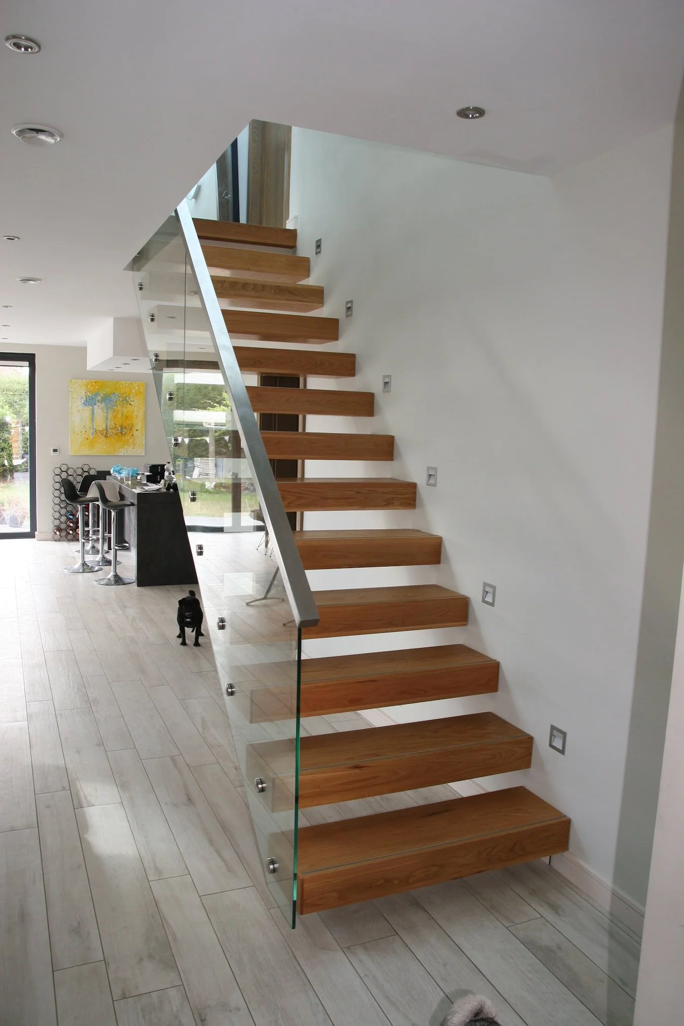 Interior of a modern house with a wooden staircase with glass railing, a black dog on the floor, and a kitchen in the background with a yellow painting on the wall.