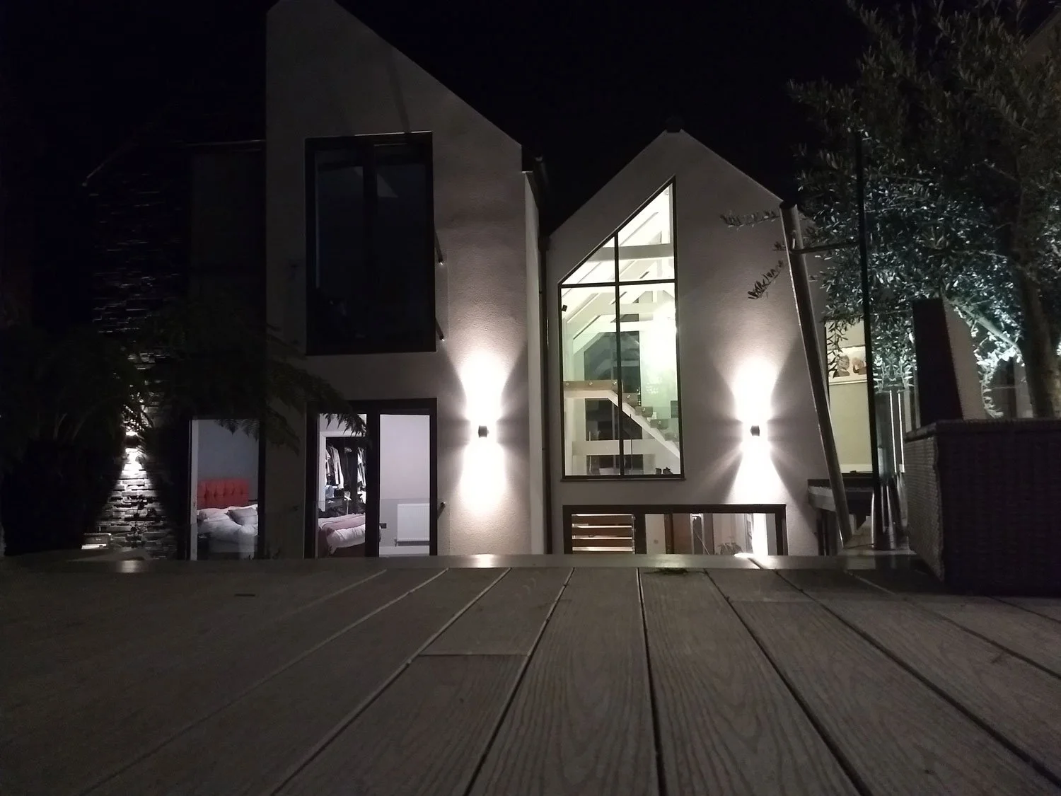 Night view of a modern house with illuminated exterior walls, large glass windows showing the interior staircase, and bedroom visible through a door on the ground floor.
