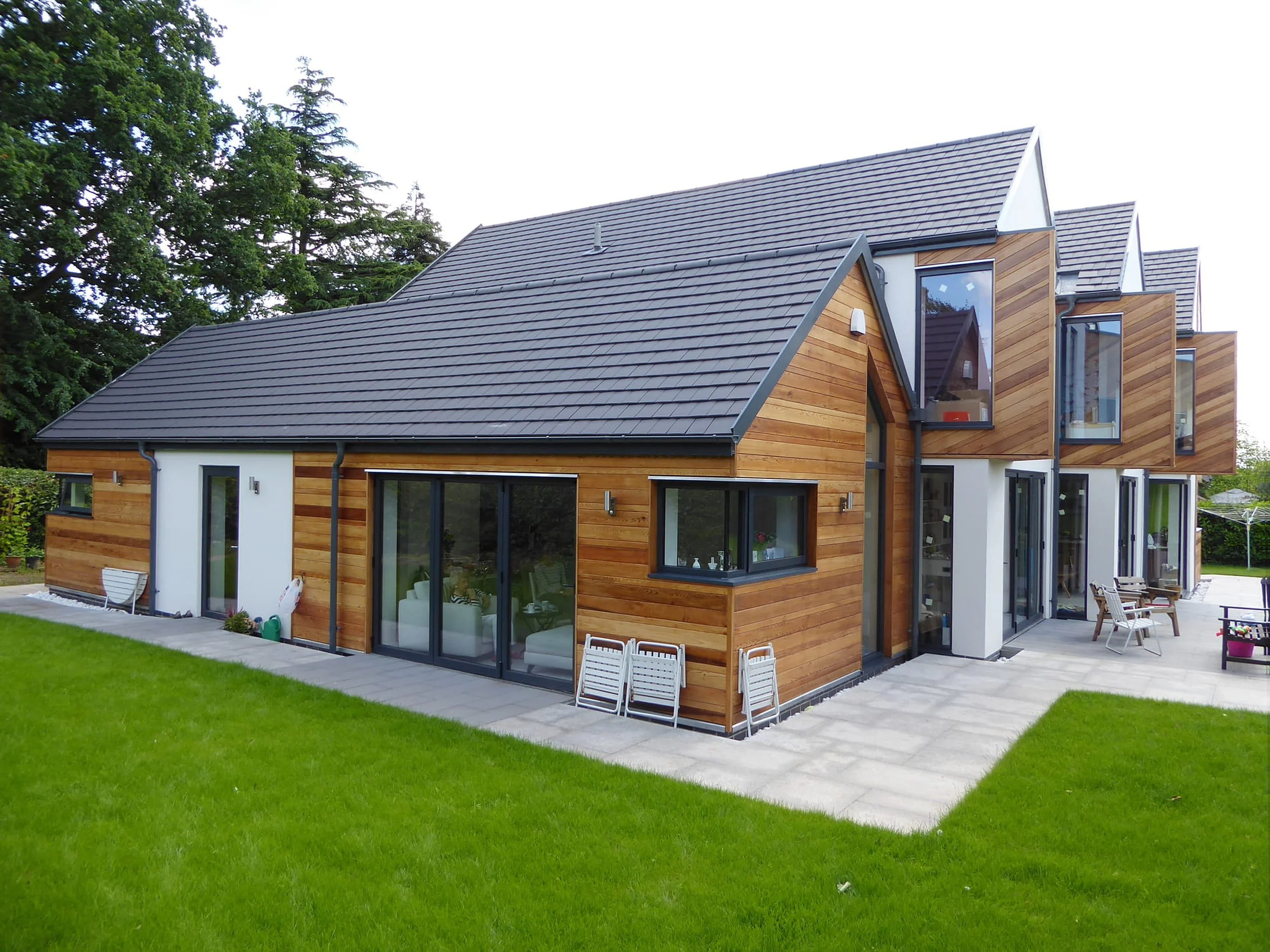 Modern house with wooden exterior and large glass windows, surrounded by green lawn and trees, with patio furniture outside.