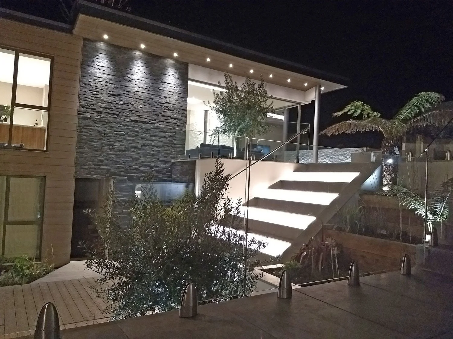 Night view of a modern house with illuminated outdoor stairs, glass railing, stone accent wall, and lush plants, including a large fern.