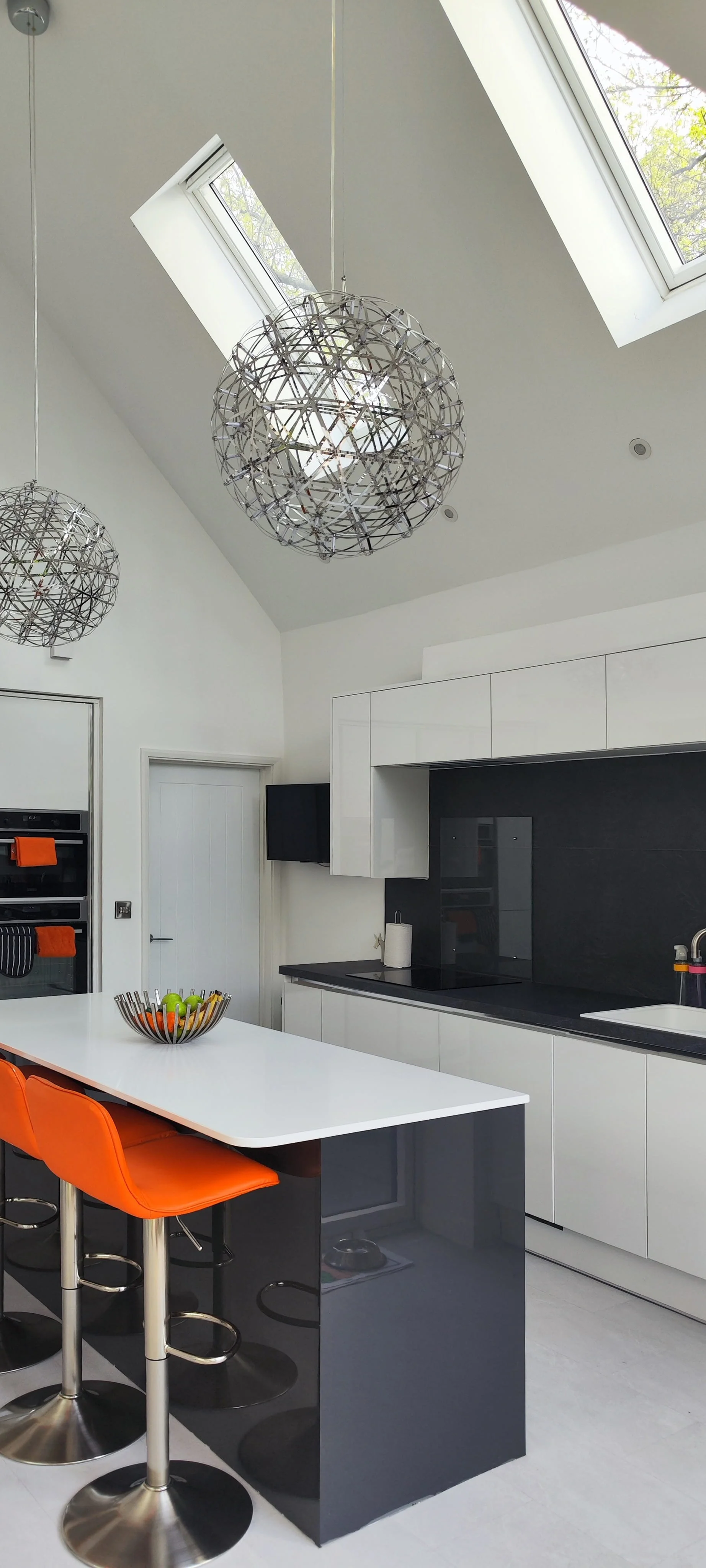 Modern kitchen with white cabinets, black countertop, and black backsplash. Two spherical pendant lights hang from a ceiling with skylights, and orange bar stools sit at a white kitchen island with a fruit bowl.