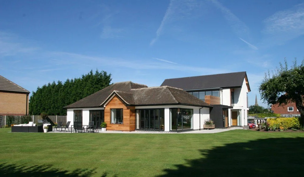 Modern house with white and wood exterior, large windows, surrounded by a lush green lawn and trees, under a blue sky.