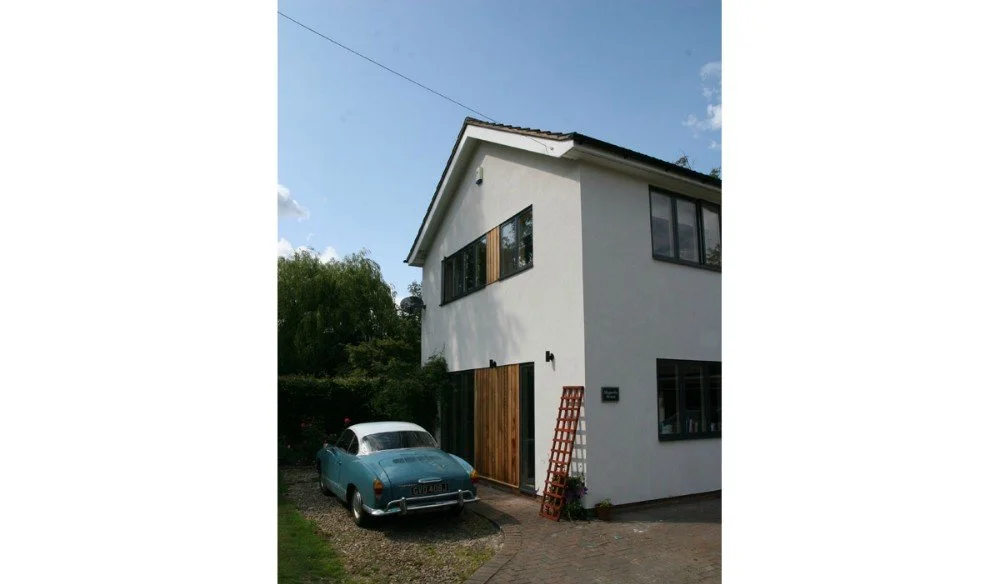 A modern white two-story house with dark window frames and a wooden front door. A vintage blue car is parked in the gravel driveway, with a small flowerpot and a lattice leaning against the house near the entrance.