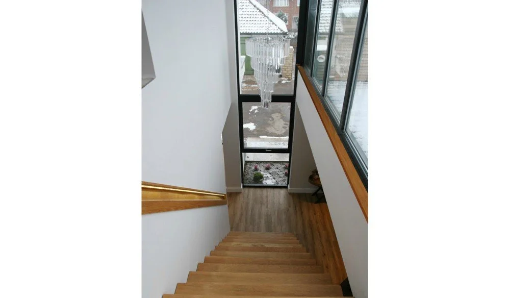Photo of a staircase from the top looking down, with a chandelier hanging overhead. The staircase has wooden steps and a wooden handrail, and it leads to a door with glass panels and a winter scene outside.
