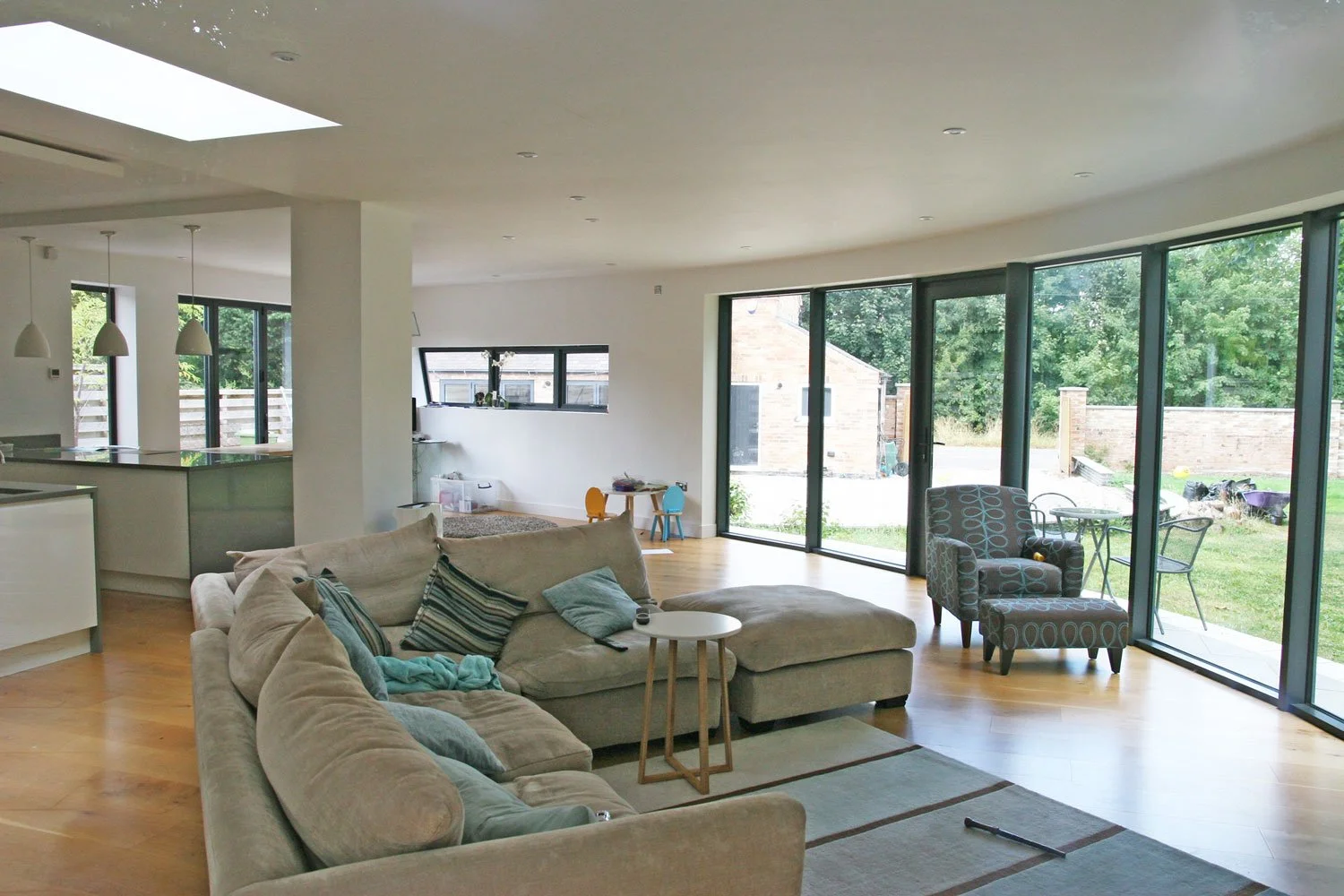 Spacious living room with large windows, beige sectional sofa with pillows, patterned armchair, small round side table, wooden flooring, and a view of the garden outside.