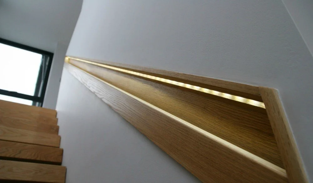 Close-up of a wooden handrail with a hidden LED strip light underneath, mounted on a white wall in a modern interior space.