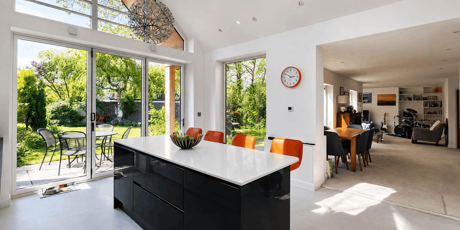 Modern open-concept kitchen and dining area with large glass sliding doors opening to a garden patio, featuring a white tabletop island, orange chairs, and a view of a lush green backyard with trees and outdoor dining furniture.