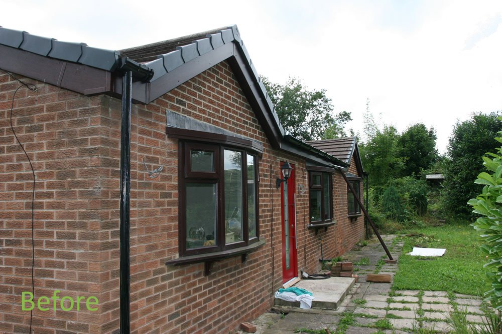 Side view of a brick house with a red door, black window frames, and a sloped roof, with a yard and greenery in the background.