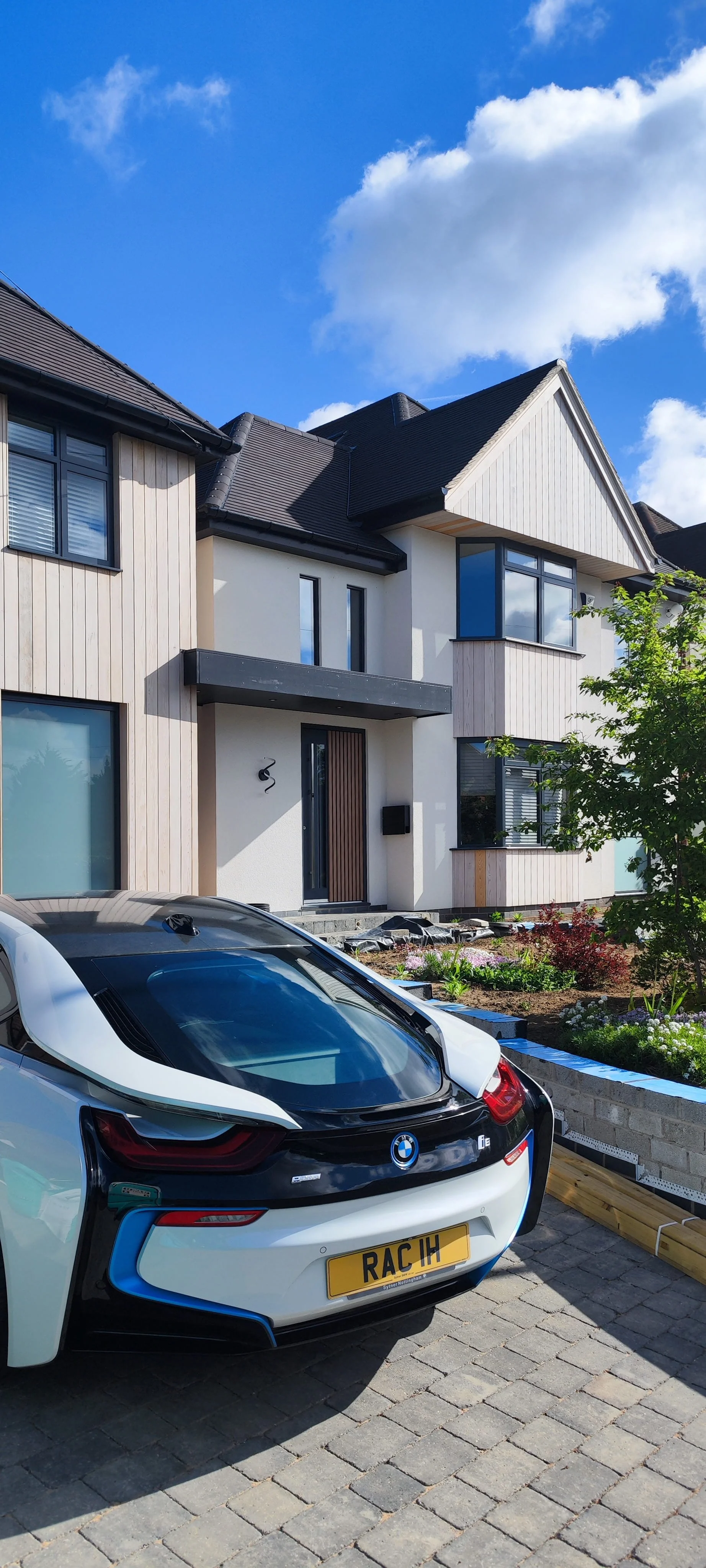 Modern house with a white and wood exterior, large windows, and a black roof. A white BMW i8 with a custom license plate 'RAC IH' is parked in front, and there is a garden with flowers and plants.