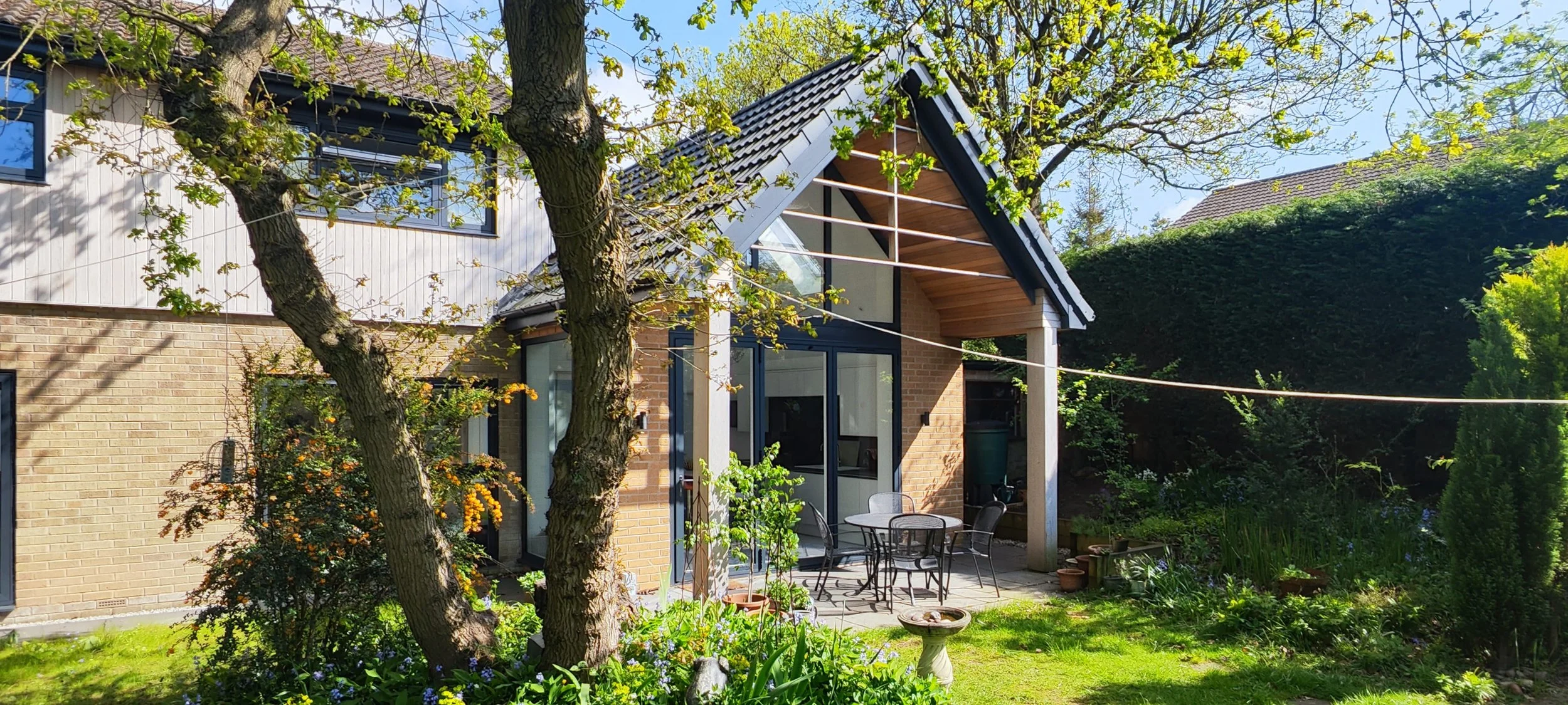 A backyard scene with a modern house, outdoor table with chairs, garden, trees, and clothesline with a sunny sky.