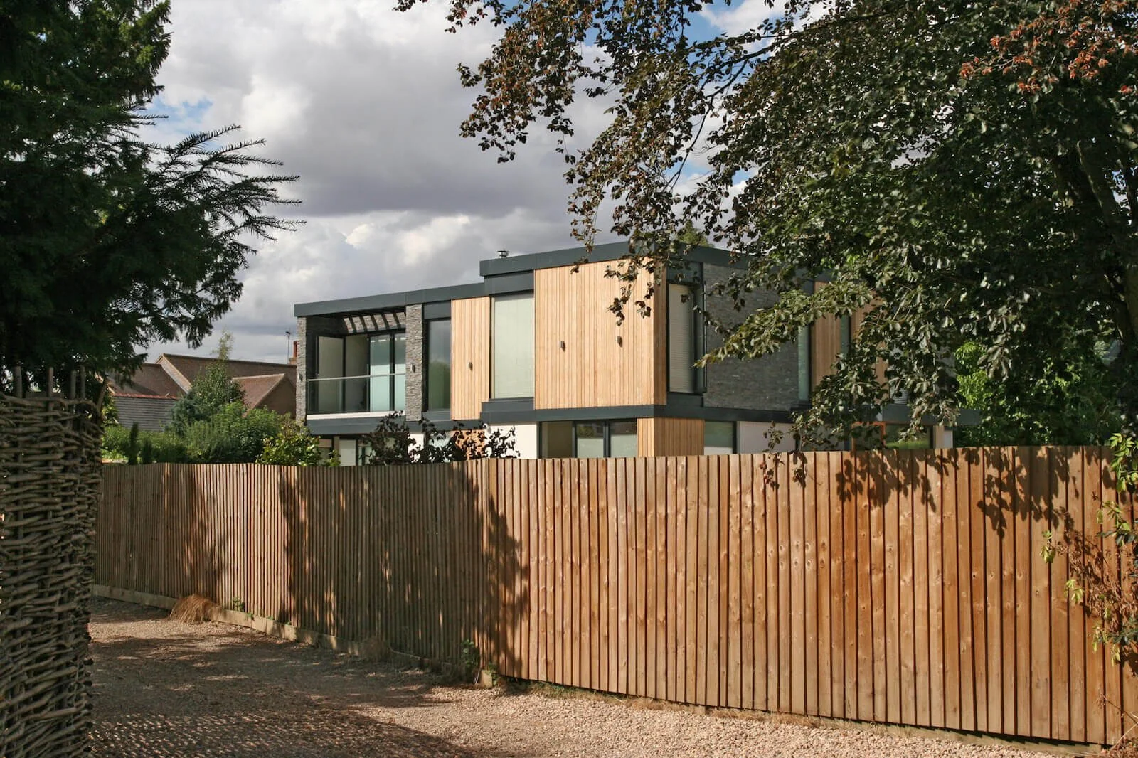 Modern house with large windows and wooden exterior panels, surrounded by trees and a wooden fence under partly cloudy sky.