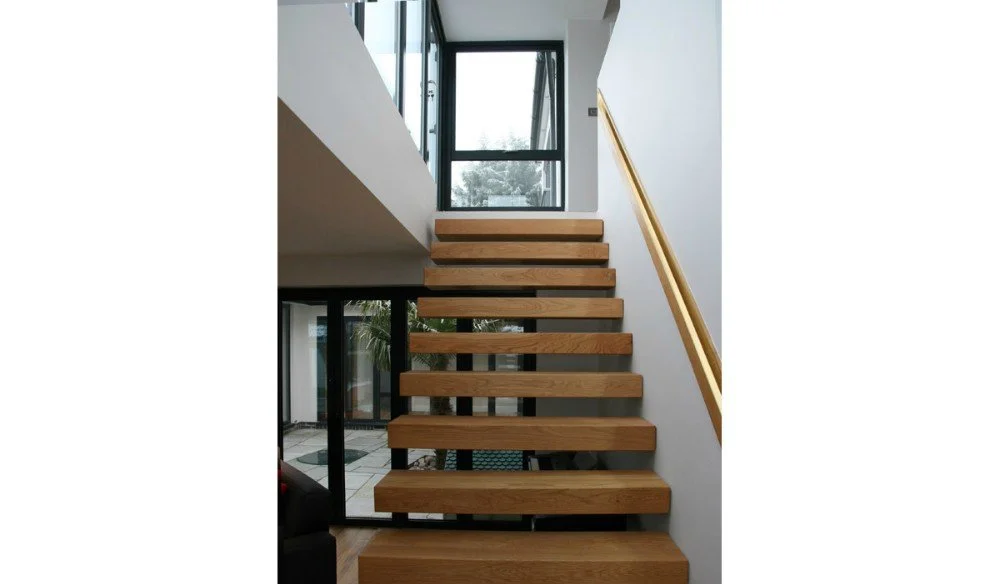 Wooden staircase with a gold handrail leading up to a large window in a modern home.