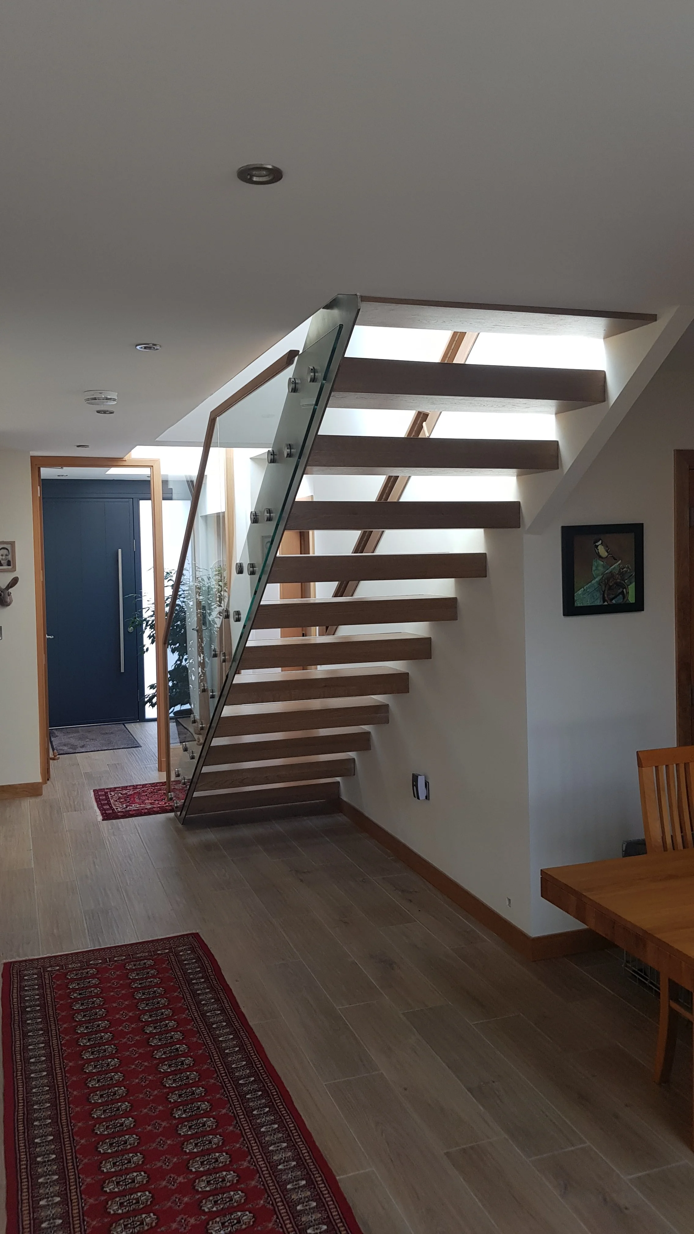Modern interior home entrance with staircase, wooden treads, glass railing, front door with a small rug, hardwood flooring, and artwork on the wall.