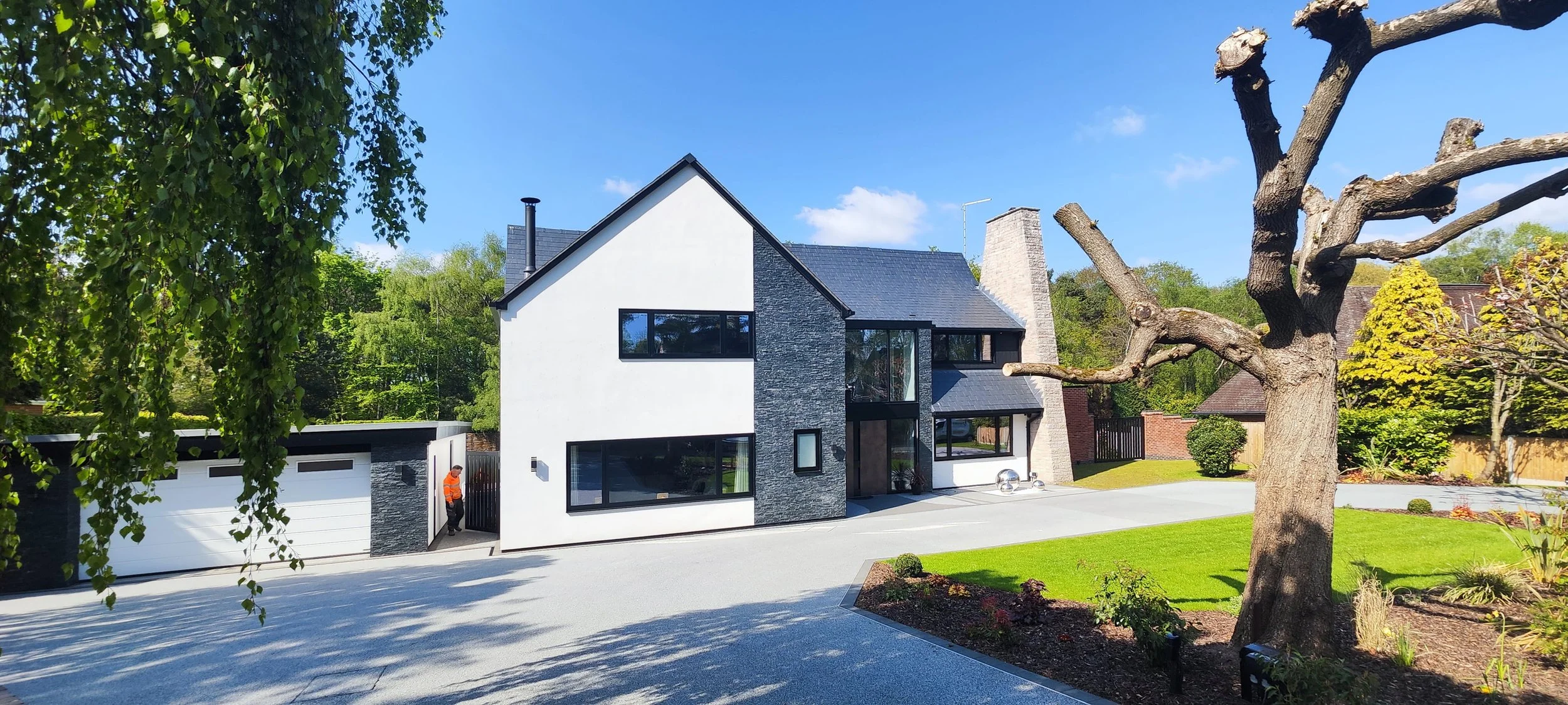 Modern white house with black-framed windows, a stone chimney, and a black garage door surrounded by green trees, a neatly maintained lawn, and a clear blue sky.