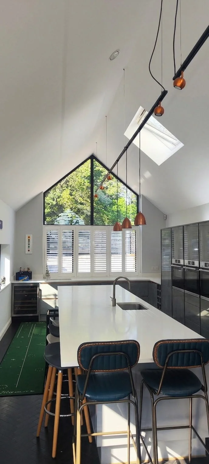 Modern kitchen with large central island, black cabinetry, and a bright triangular window with green trees outside. Pendant lights hang above the island, and there's a skylight in the ceiling.