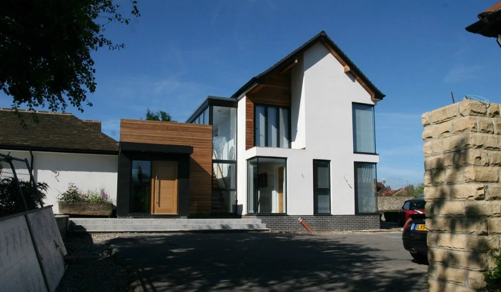 A modern two-story house with a white exterior, large windows, and a mix of wood and black accents, situated on a paved lot under a blue sky.