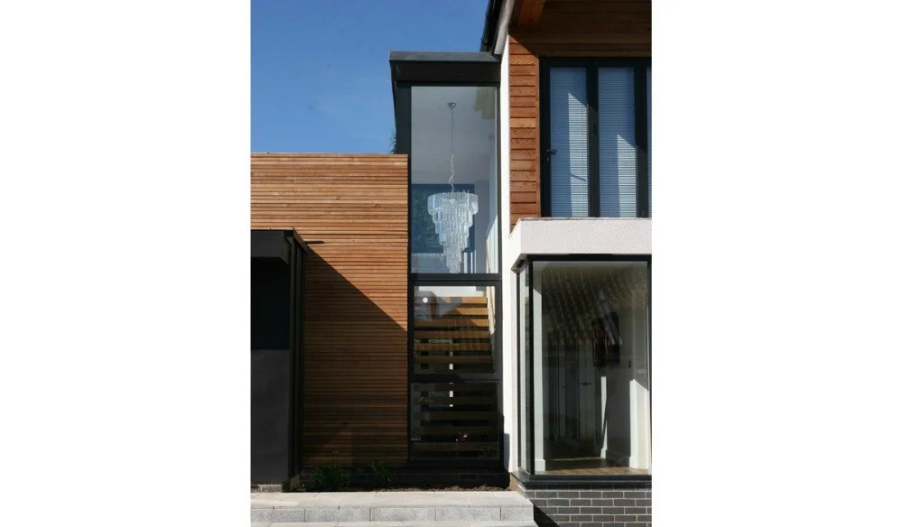 Modern house with a mix of wood, glass, and brick exterior, featuring large windows and a chandelier visible through the glass.