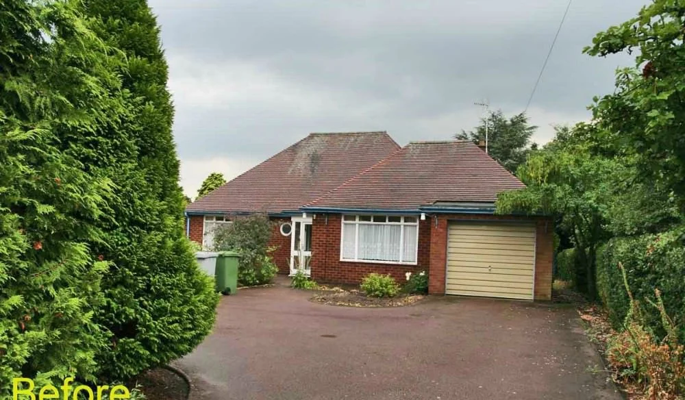A single-story brick house with a sloped roof, a garage on the right, and a driveway, surrounded by tall green trees, under a cloudy sky.