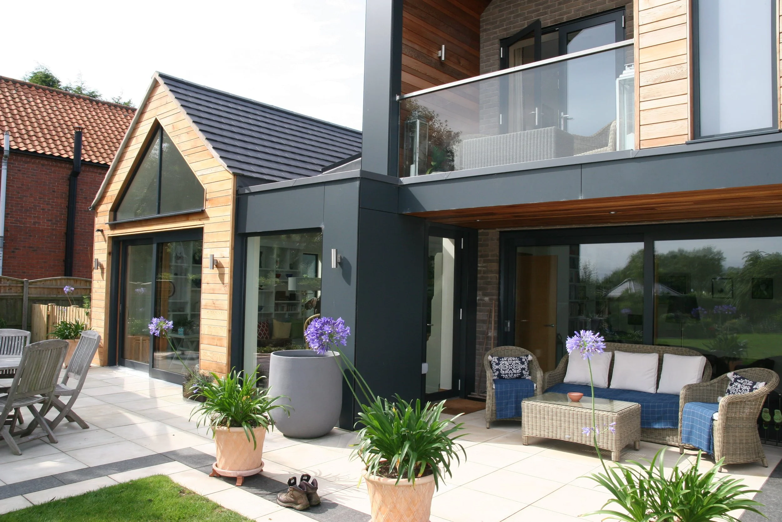 Modern house with a balcony and patio area, outdoor furniture, potted plants, and a garden with purple flowers.