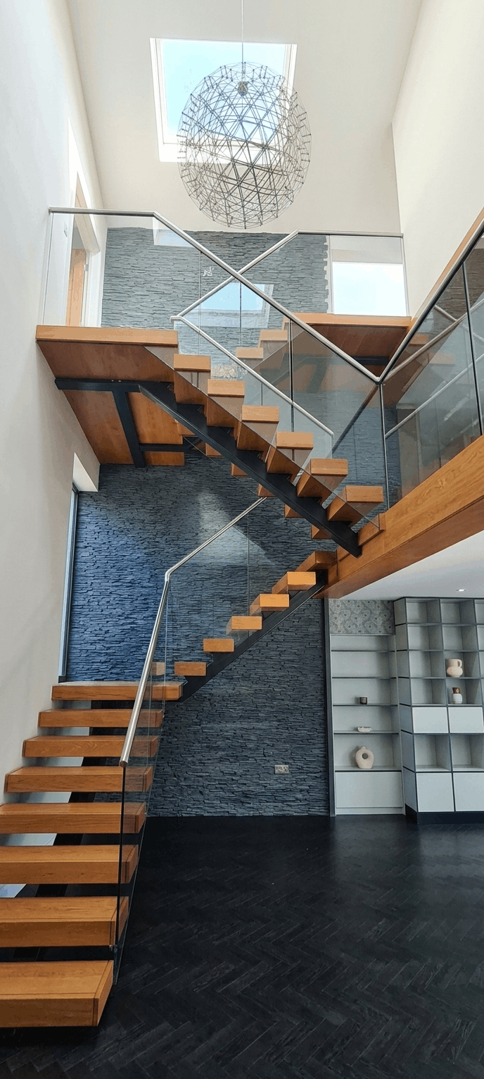Modern interior staircase with wooden steps, glass railing, black metal supports, gray stone wall, white shelving unit, and a spherical metal chandelier hanging from the ceiling near a skylight.
