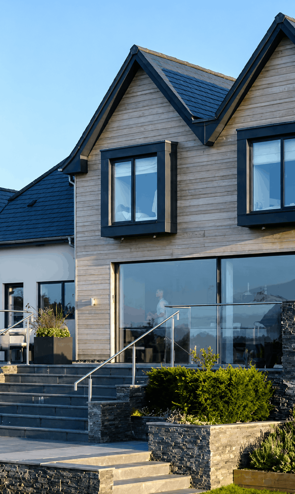 Modern house with wooden siding, black-framed windows, glass sliding doors, and landscaped steps with green bushes.