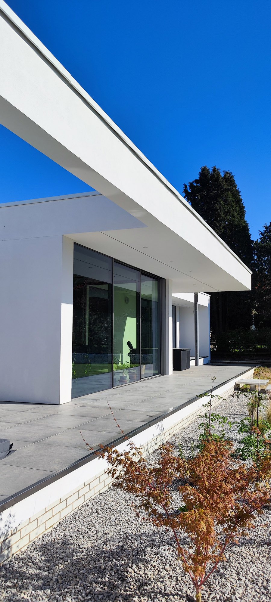 Modern white house with large glass doors, a flat patio, and a gravel garden with a small reddish-brown tree under a clear blue sky.
