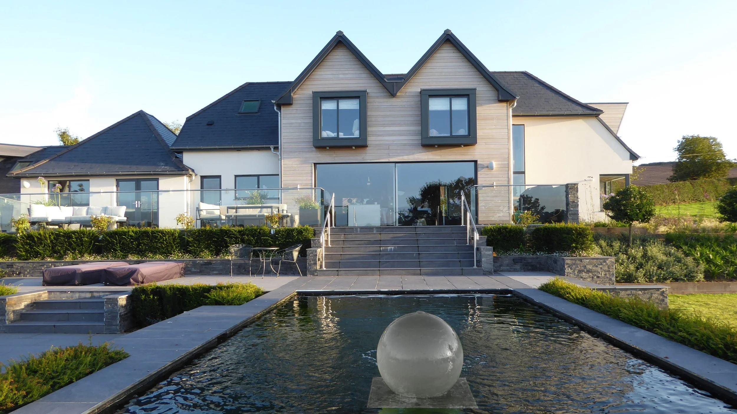 Modern house with a patio, stairs, and a water feature in the foreground, surrounded by greenery and outdoor furniture.