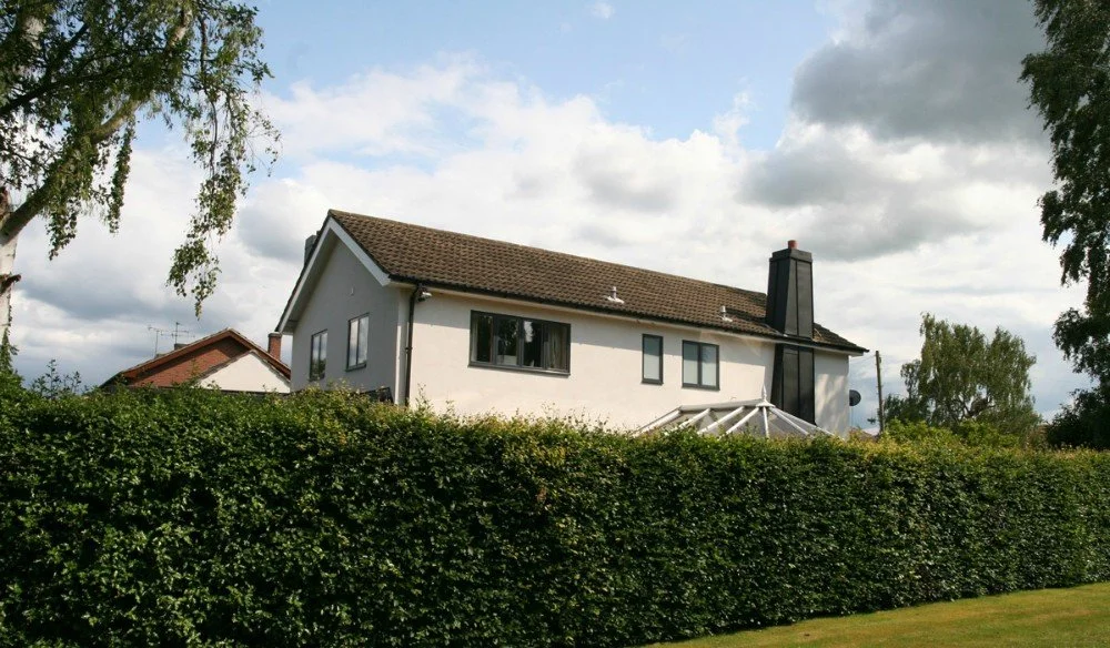 A white two-story house with a brown roof, black chimney, and a glass porch, surrounded by a tall green hedge and trees, under a partly cloudy sky.