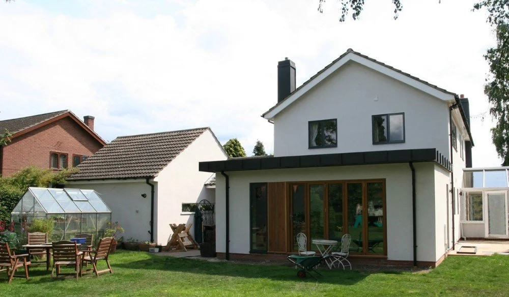 A two-story white house with a gabled roof and black accents, a glass patio door, and outdoor furniture on a grassy yard with a small greenhouse and another house in the background.