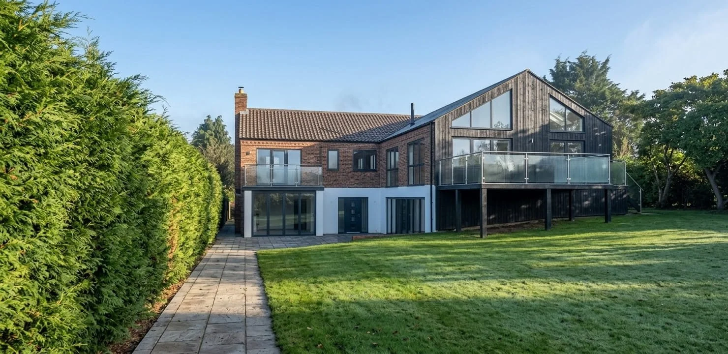 Modern two-story house with a mix of brick and dark wood exterior, large windows, glass balconies, and a spacious grassy backyard.