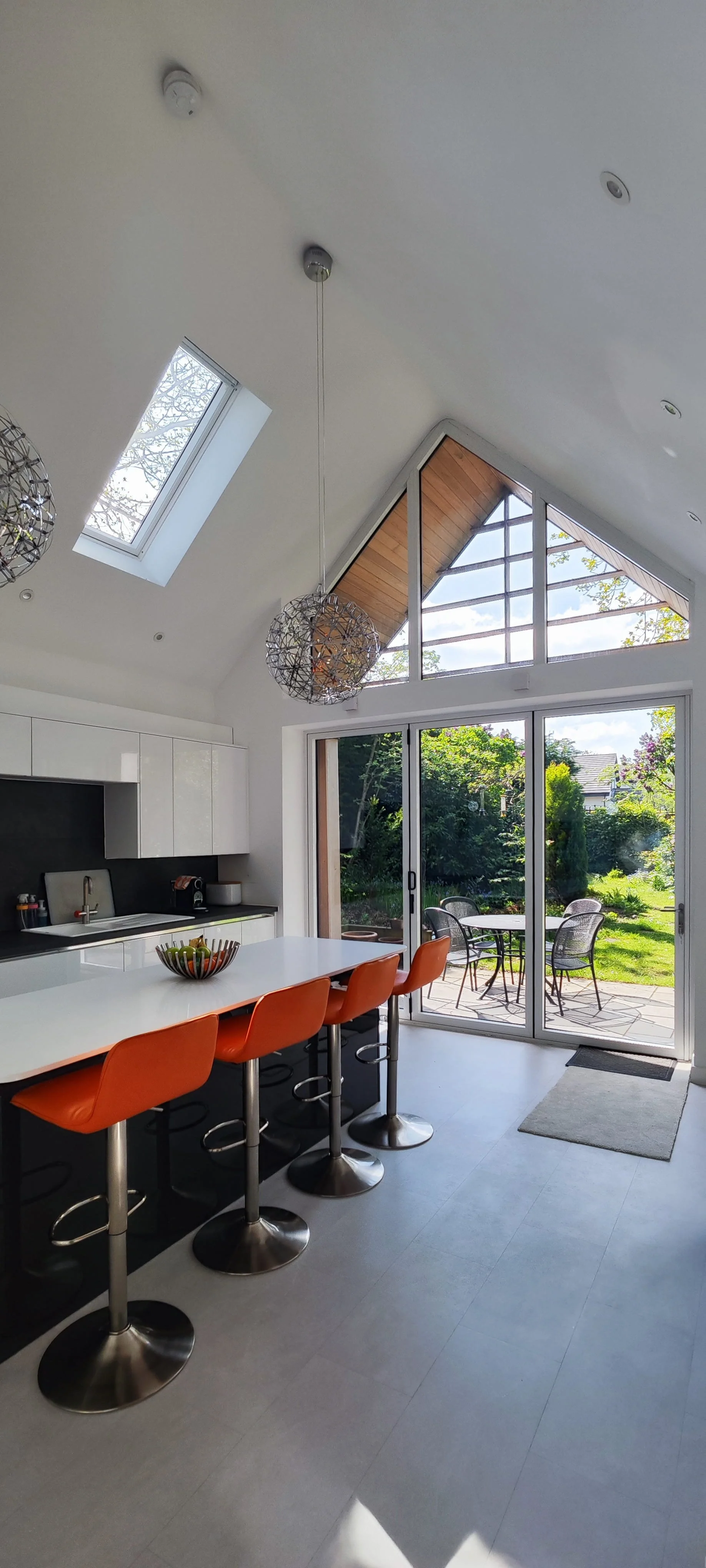 Modern kitchen with large glass sliding doors leading to a garden patio, featuring a white island with orange bar stools, white cabinets, black backsplash, and two spherical pendant lights hanging from a high vaulted ceiling with skylights.
