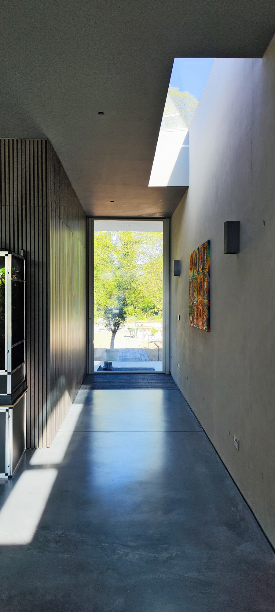 A modern hallway with a large glass door at the end, outside trees and a bench visible. The right wall is decorated with a colorful abstract painting, and there are black wall sconces. The left side features a wooden slat wall and what appears to be 