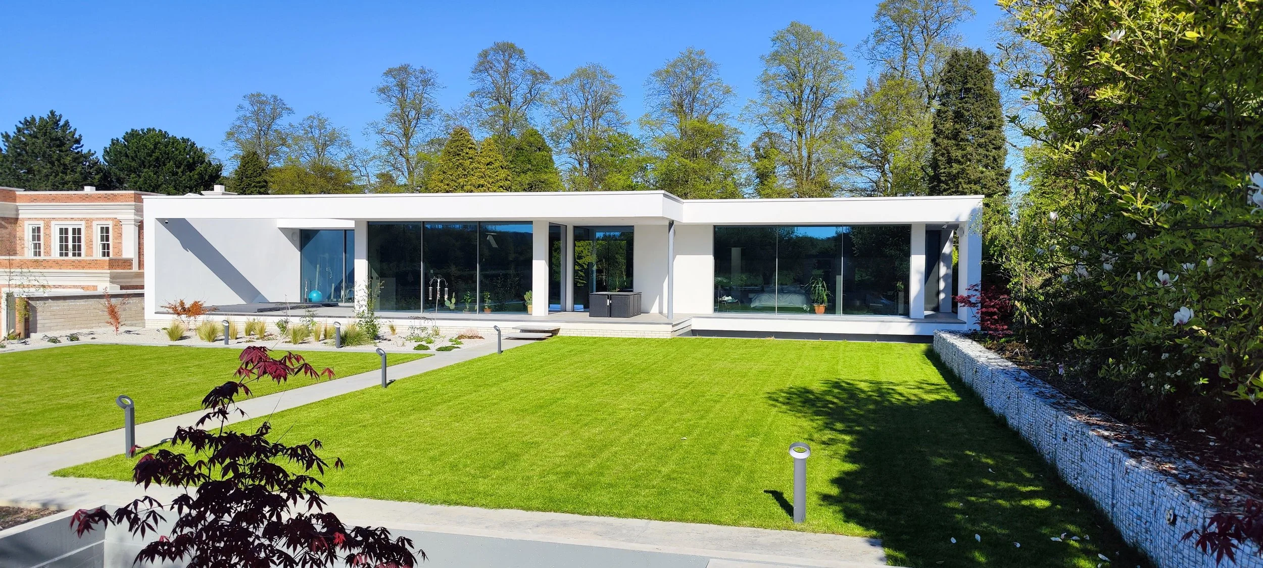 Modern white house with large glass windows and a well-maintained green lawn, trees in the background, and a clear blue sky.
