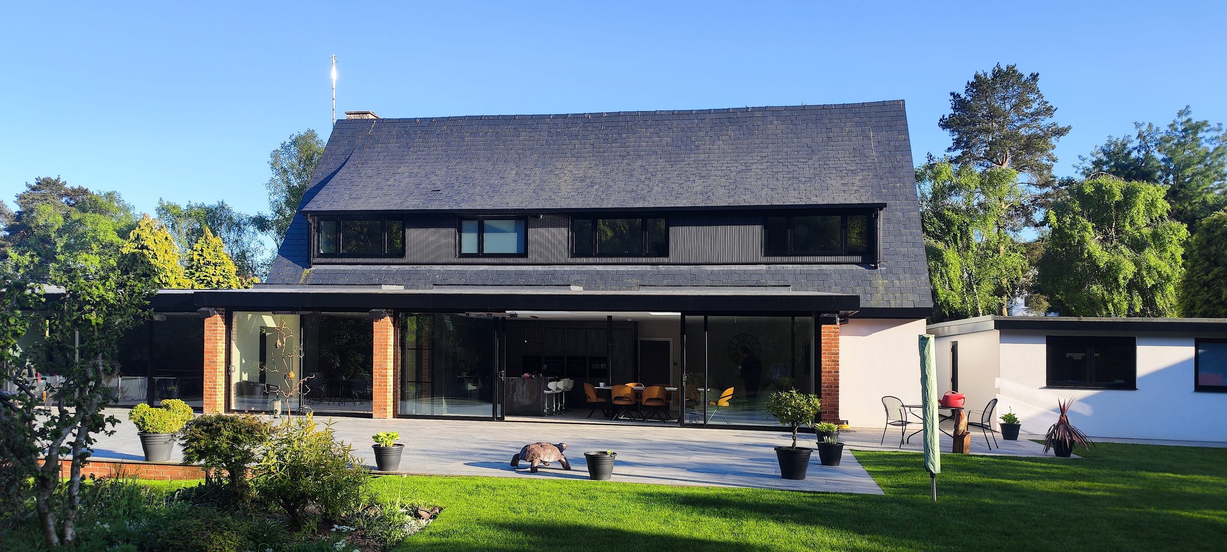 A modern two-story house with a black shingled roof, large sliding glass doors, and a patio with potted plants and outdoor furniture, surrounded by a green lawn and trees.