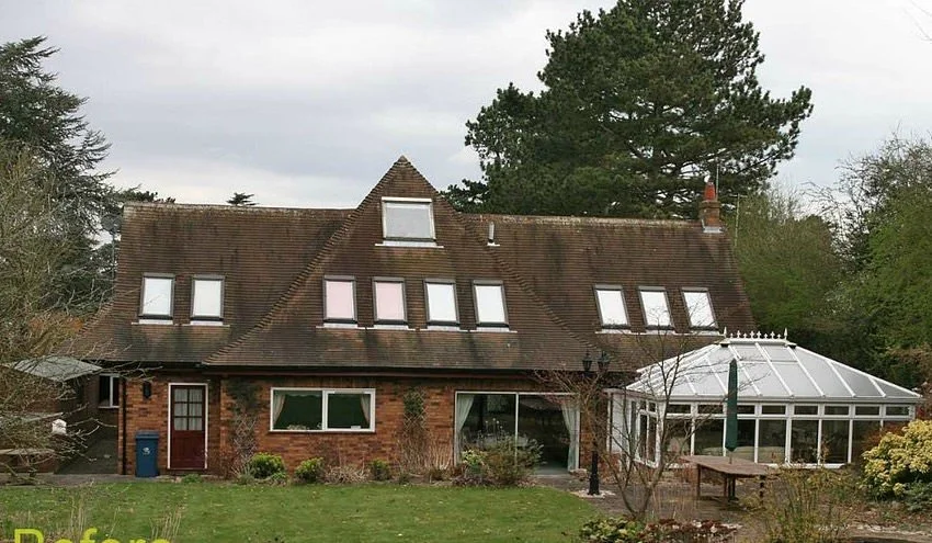 Brick house with multiple dormer windows and a glass conservatory attached on the right side, surrounded by trees and a grassy yard.