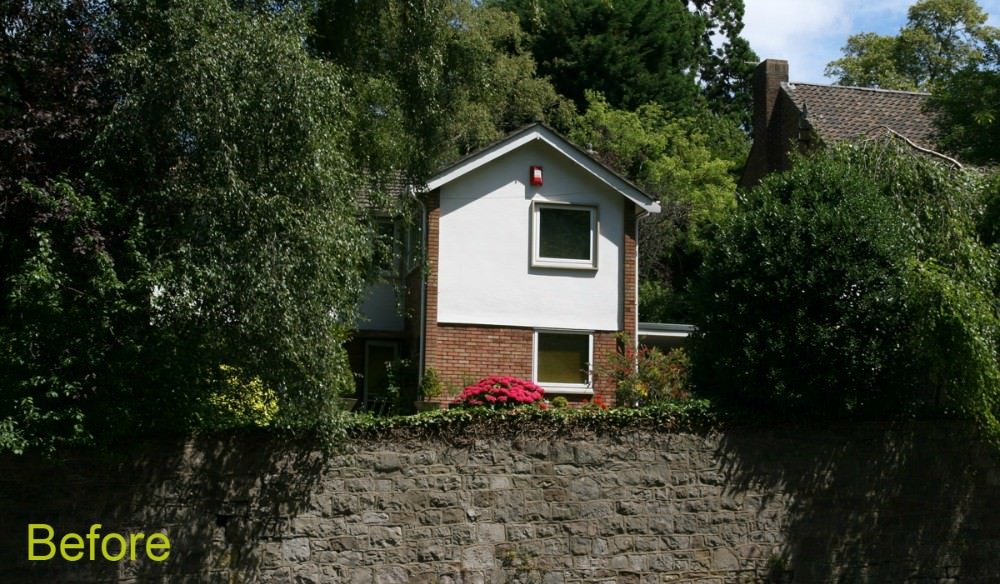 A two-story house with white siding on the upper level and brick on the lower level, surrounded by trees, with a stone retaining wall in the foreground and a small lawn with pink flowers in front.