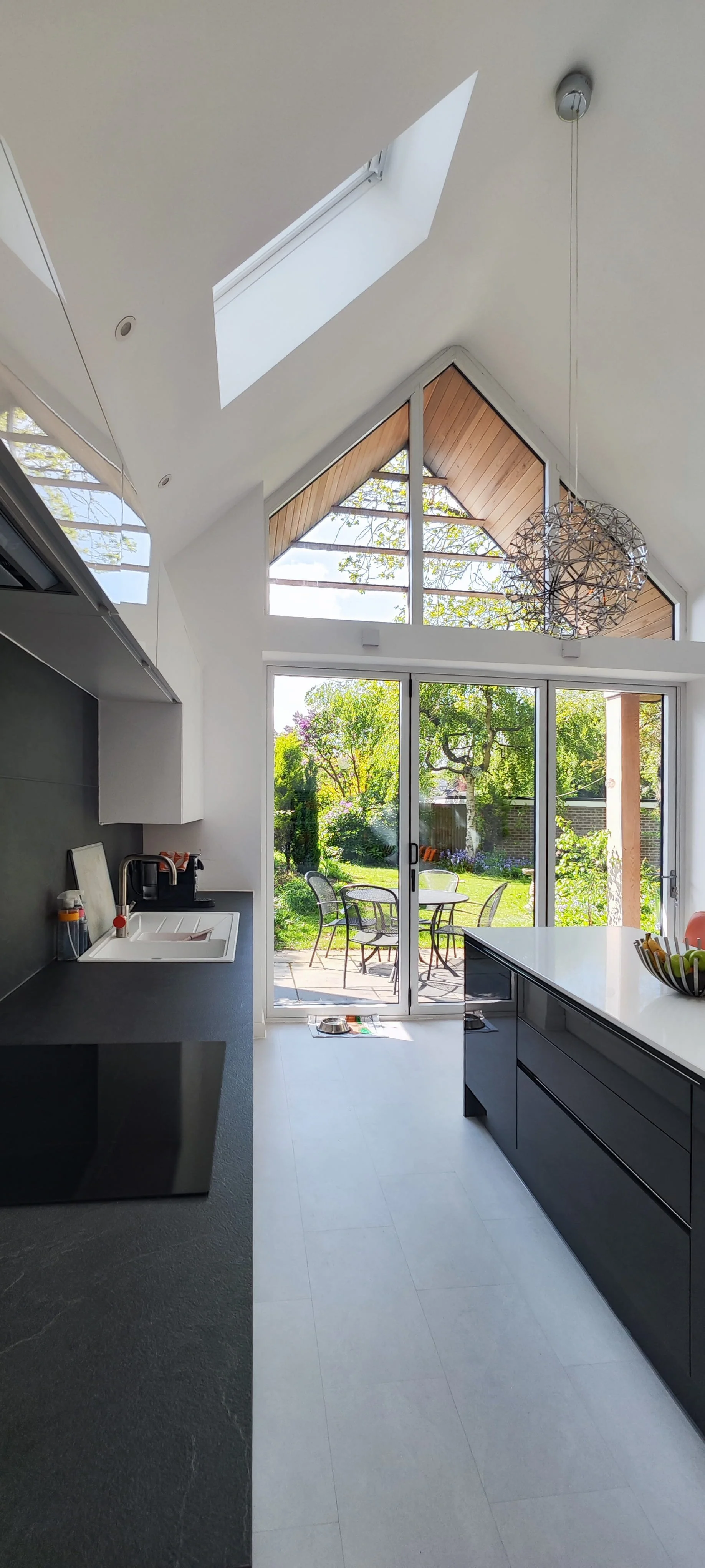 Modern kitchen with large sliding glass door opening to a lush backyard patio with trees and outdoor furniture, ceiling skylights, and contemporary pendant lighting.