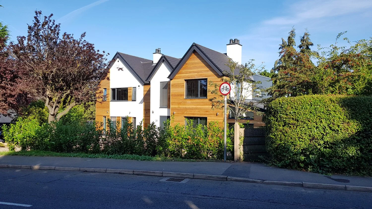 Modern residential buildings with wooden and white exteriors, front garden with bushes and trees, street sign indicating no bicycles, blue sky, and a paved curb.